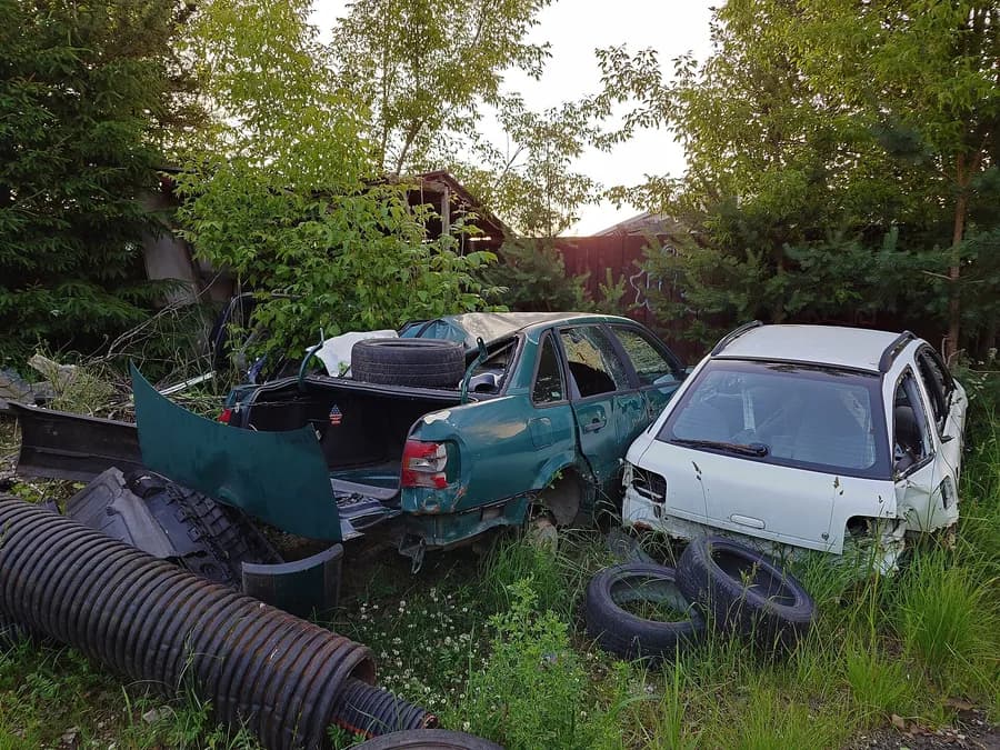 Açık alanda yan yana duran, camları kırılmış ve parçaları sökülmüş yeşil sedan ile beyaz hatchback hurda araçlar