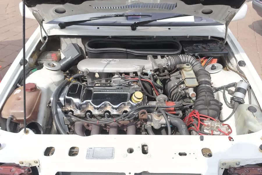 Motor kaputu açılmış beyaz otomobilin yıpranmış motor bölümü; silindir kapağı, bujiler ve hortumlar üstten görünüyor