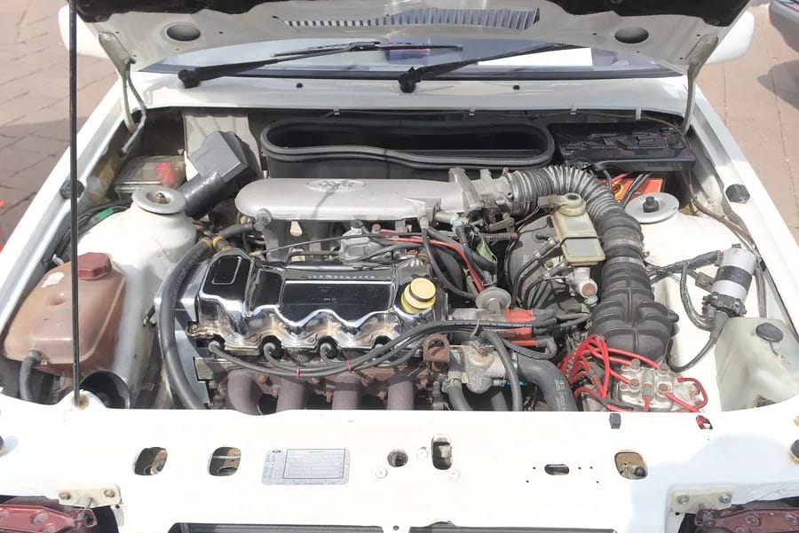 Motor kaputu açılmış beyaz otomobilin yıpranmış motor bölümü; silindir kapağı, bujiler ve hortumlar üstten görünüyor